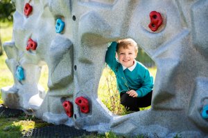 a child smiling and peeking through a hole of a climbing wall outside